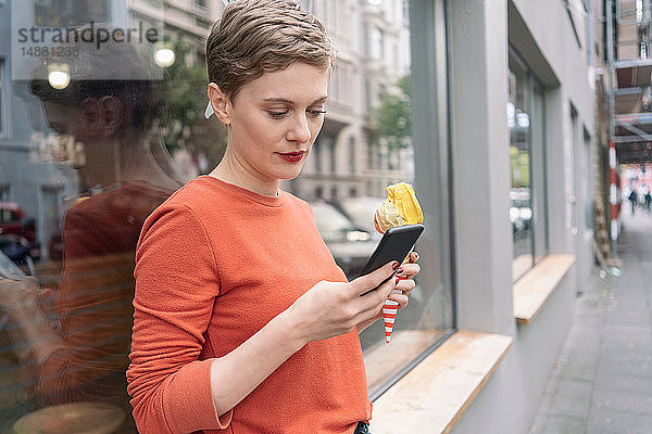 Frau hält Eiscreme in der Hand und benutzt ein Smartphone vor dem Geschäft  Köln  Nordrhein-Westfalen  Deutschland