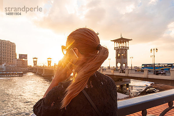 Touristin mit roten Haaren beim Smartphone-Anruf in der Nähe der Stanley-Brücke bei Sonnenuntergang  Rückansicht  Alexandria  Ägypten