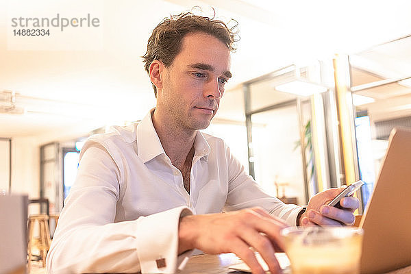 Mittlerer erwachsener Geschäftsmann  der ein Smartphone betrachtet  während er einen Laptop am Bürotisch benutzt