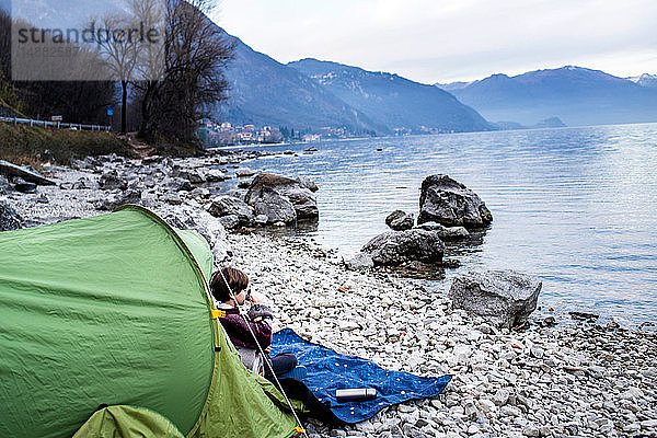 Junge sitzt auf einer Decke am Zelt und trinkt Kaffee am Seeufer  Comer See  Onno  Lombardei  Italien