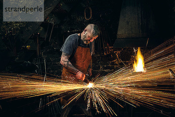 Ein reifer männlicher Schmied macht Funken  indem er in der Werkstatt Metall auf den Amboss hämmert