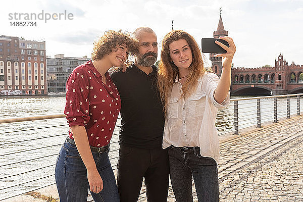 Mann und Freundinnen beim Selbstfahren mit dem Smartphone auf Brücke  Fluss und Gebäuden im Hintergrund  Berlin  Deutschland