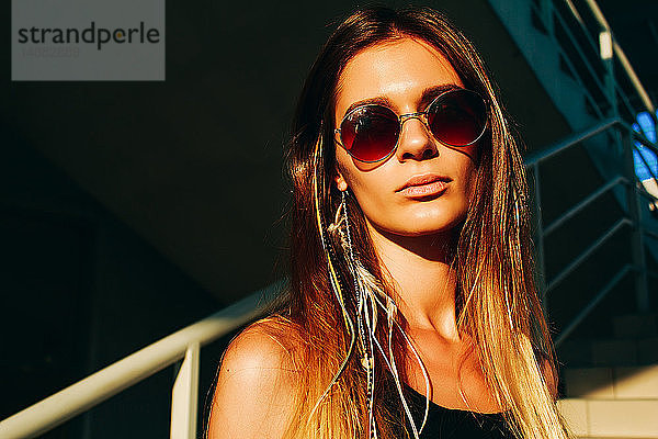 Junge Frau mit langen braunen Haaren und Sonnenbrille auf Treppen-  Kopf- und Schulterportrait