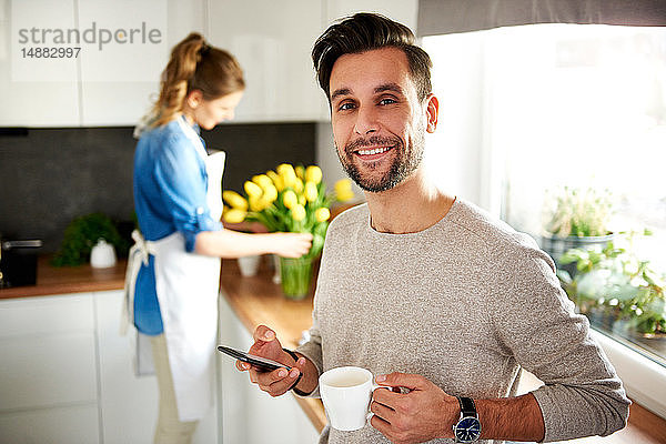 Mann benutzt Smartphone in der Küche  Frau arrangiert Blumen im Hintergrund