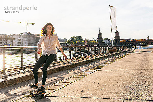 Junge Frau beim Skateboarden auf Brücke  Fluss und Gebäuden im Hintergrund  Berlin  Deutschland