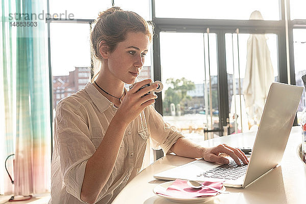 Junge Frau benutzt Laptop und trinkt an sonnigen Tagen Kaffee im Büro
