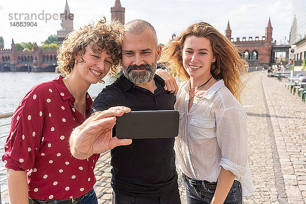Mann und Freundinnen beim Selbstfahren mit dem Smartphone auf Brücke  Fluss und Gebäuden im Hintergrund  Berlin  Deutschland