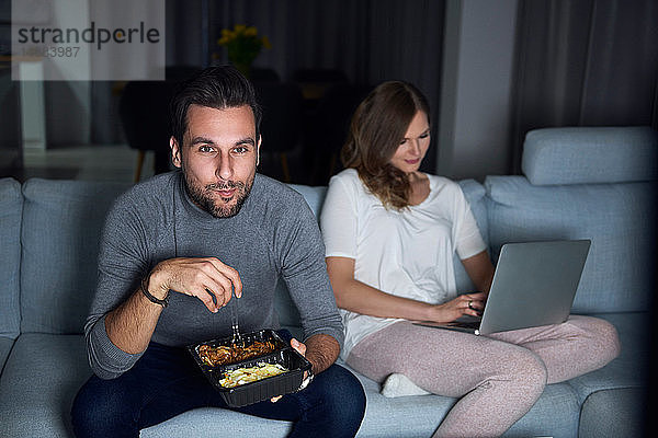 Junges Paar sitzt abends auf dem Sofa  isst zum Mitnehmen und benutzt Laptop