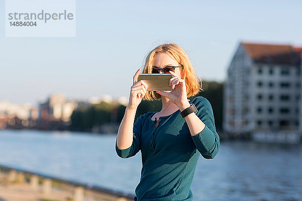 Junge Frau fotografiert mit einem Smartphone am Fluss im Sommer  Berlin  Deutschland
