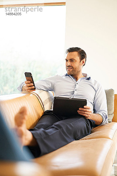 Reifer Mann schaut auf Smartphone  während er auf dem Sofa liegt