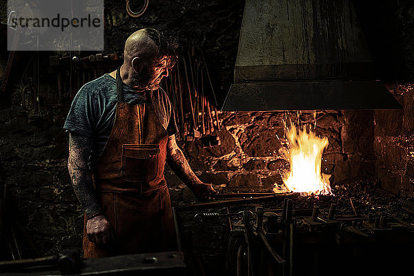 Ein reifer männlicher Schmied erhitzt Metall auf offenem Feuer in einer Werkstatt
