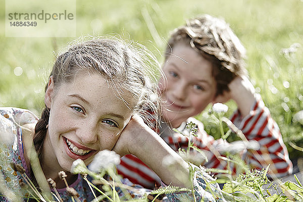 Porträt eines lächelnden Mädchens auf einer Wiese liegend mit kleinem Bruder im Hintergrund