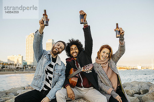 Porträt von drei glücklichen Freunden mit Gitarre  die bei Sonnenuntergang an der Küste Bierflaschen hochheben