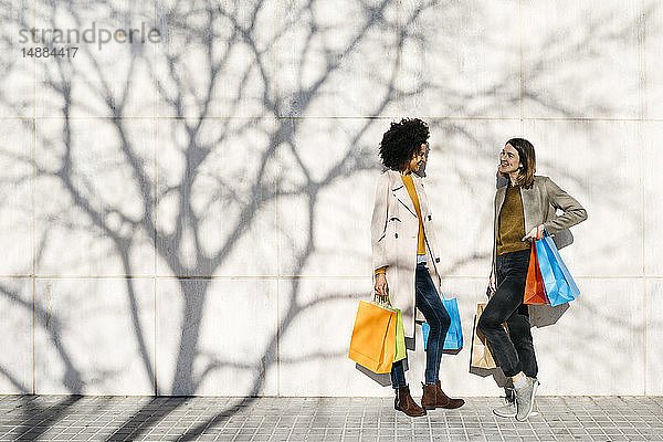 Zwei glückliche Frauen mit Einkaufstaschen stehen an einer Wand und unterhalten sich