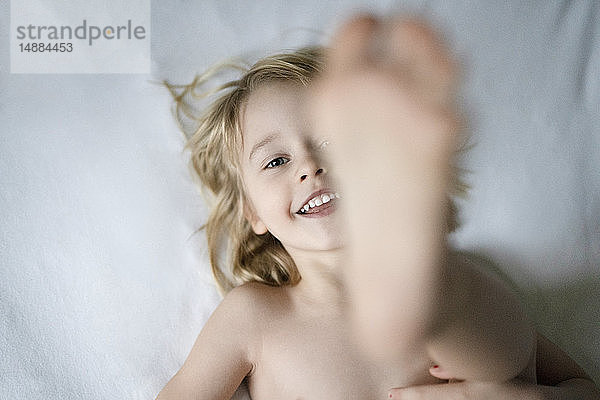 Porträt eines lächelnden blonden Mädchens  das auf einem Bett liegt und ihre Füße zeigt  Draufsicht