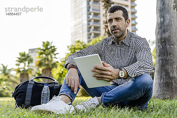 Porträt eines Mannes mit digitalem Tablett  der auf einer Wiese im Stadtpark sitzt und nachdenkt