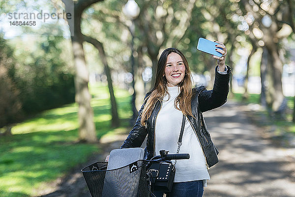 Lächelnde junge Frau mit Fahrrad im Park  die ein Selfie nimmt