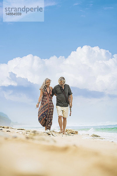 Älteres Hippie-Paar mit Gitarre Arm in Arm am Strand spazieren