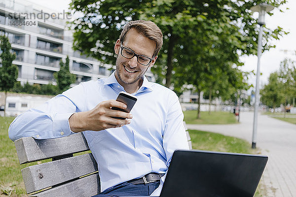 Junger Geschäftsmann auf Parkbank sitzend  mit Smartphone und Laptop