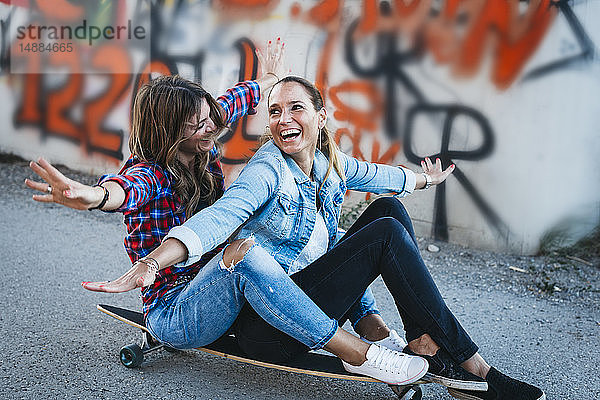 Zwei lachende Freunde sitzen mit ausgestreckten Armen auf einem Longboard