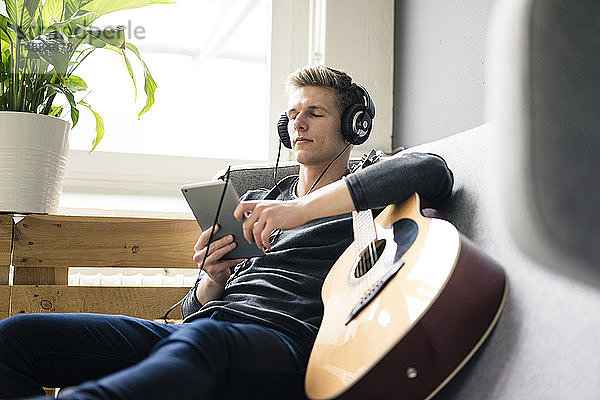 Entspannter junger Mann mit Tablett und Gitarre  der mit Kopfhörern Musik hört