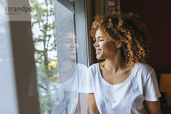 Lächelnde junge Frau mit lockigem Haar  die zu Hause aus dem Fenster schaut