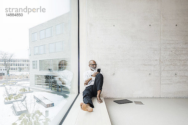 Barfüßiger reifer Geschäftsmann sitzt auf Fensterbank mit Smartphone