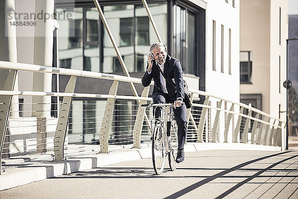 Lächelnder reifer Geschäftsmann mit Kopfhörern beim Fahrradfahren auf einer Brücke in der Stadt