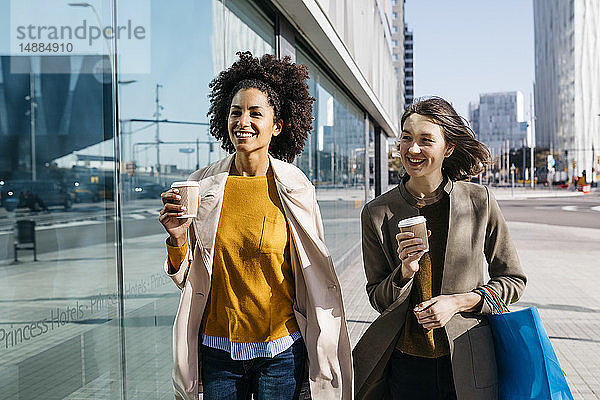 Zwei glückliche Frauen mit Einkaufstaschen und Kaffee zum Mitnehmen spazieren durch die Stadt