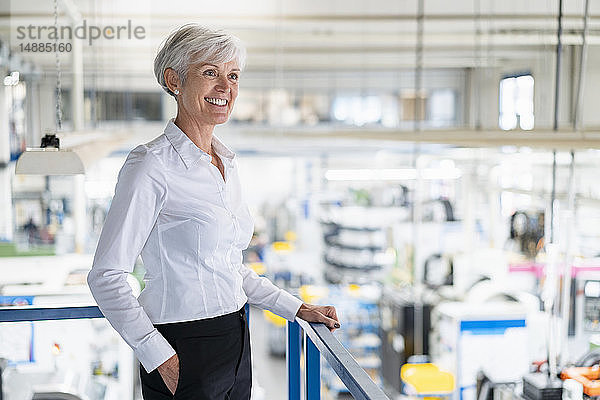 Lächelnde ältere Geschäftsfrau im oberen Stockwerk einer Fabrik mit Blick auf die Fabrikhalle