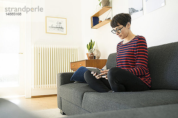 Lässige kurzhaarige Frau  die sich auf einer Lounge-Couch im modernen Wohnzimmer mit Hilfe eines Tablets entspannt