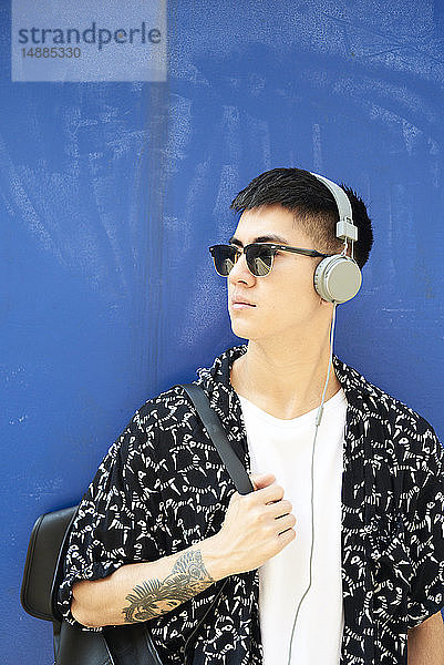 Junger Mann hört Musik mit Kopfhörern an blauer Wand