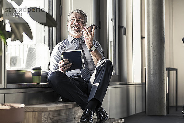 Glücklicher reifer Geschäftsmann mit Notebook  der mit seinem Handy am Fenster sitzt