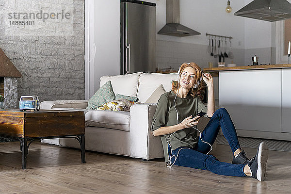 Junge Frau sitzt zu Hause auf dem Boden  benutzt ein Smartphone und trägt Kopfhörer