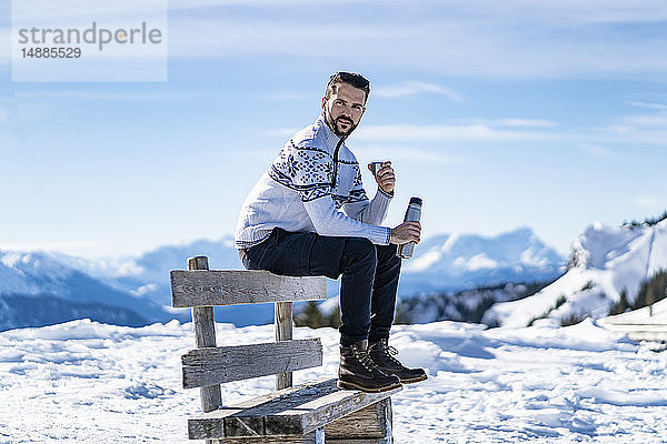 Deutschland  Bayern  Brauneck  Mann sitzt im Winter auf einer Bank in den Bergen und macht Pause