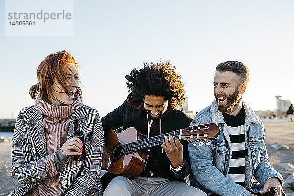 Drei glückliche Freunde mit Gitarre sitzen bei Sonnenuntergang im Freien