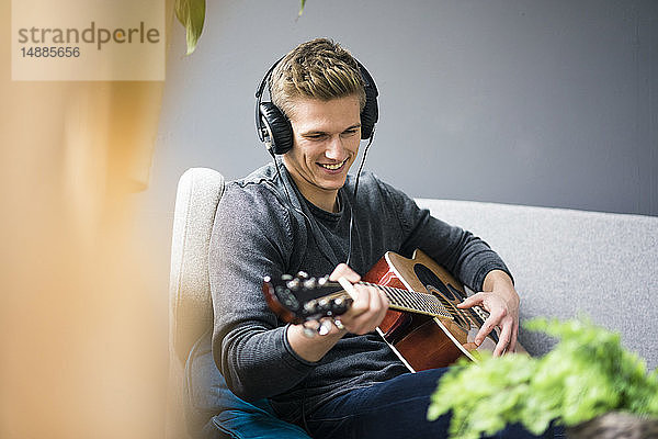 Lächelnder junger Mann mit Kopfhörern sitzt auf Couch und spielt Gitarre