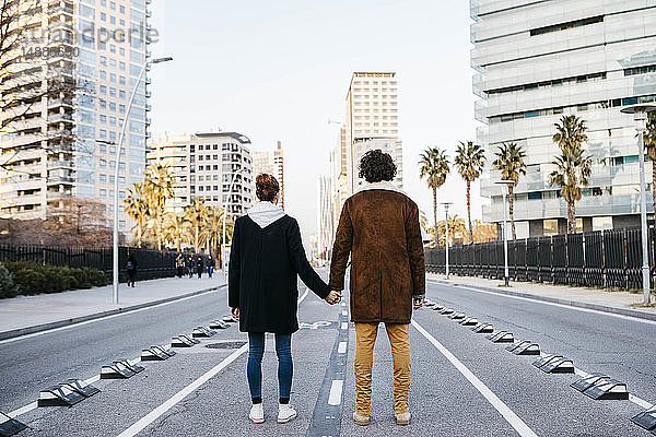Rückansicht eines Paares  das sich in der Mitte der Straße an den Händen hält und die Gebäude im Hintergrund betrachtet