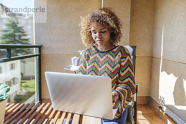 Junge Frau mit lockigem Haar sitzt mit Laptop auf dem Balkon