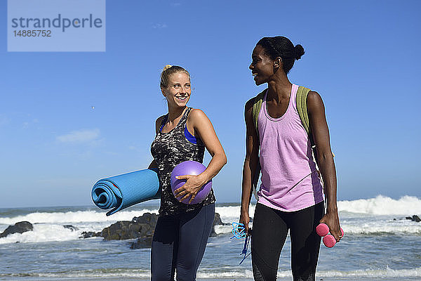 Zwei glückliche Frauen mit Fitnessgeräten am Strand spazieren