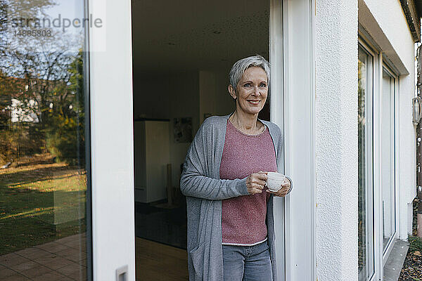 Lächelnde reife Frau mit einer Tasse Kaffee an der Terrassentür