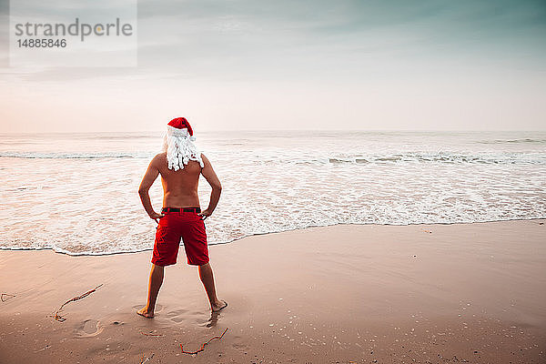 Thailand  Rückenansicht eines als Weihnachtsmann verkleideten Mannes  der bei Sonnenuntergang am Strand steht und auf den Horizont schaut