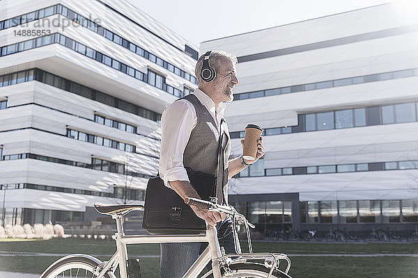 Lächelnder reifer Geschäftsmann mit Fahrrad  Kaffee zum Mitnehmen und Kopfhörern unterwegs in der Stadt