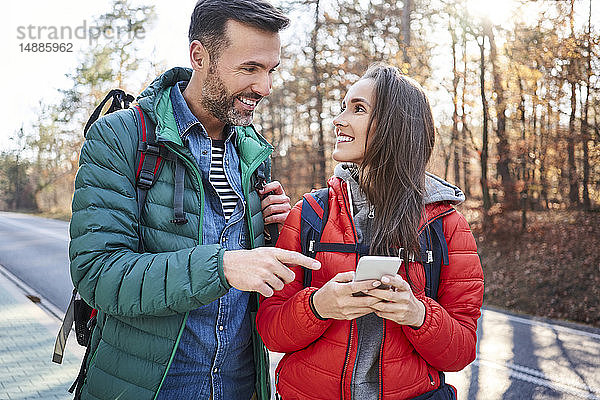 Glückliches Paar überprüft Smartphone während einer Rucksacktour auf einer Straße im Wald