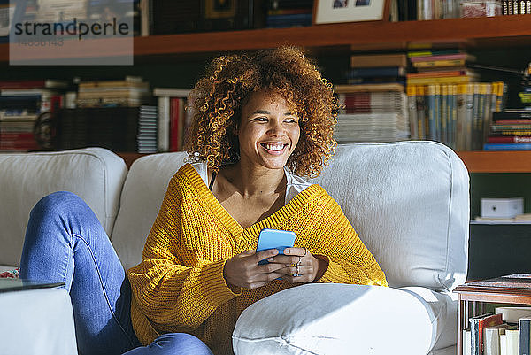 Glückliche junge Frau mit Handy sitzt zu Hause auf dem Sofa
