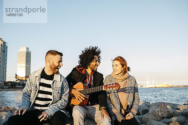 Drei glückliche Freunde mit Gitarre sitzen draußen am Sonnenuntergang an der Küste