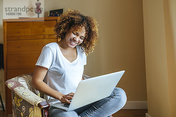 Glückliche junge Frau mit lockigem Haar sitzt zu Hause im Sessel und benutzt ihren Laptop