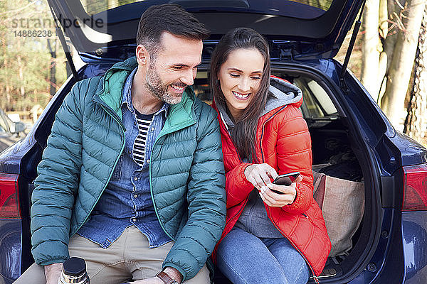 Ehepaar benutzt Smartphone  während es während einer Autofahrt im Kofferraum sitzt