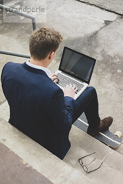 Junger Geschäftsmann mit Skateboard sitzt im Freien auf einer Treppe mit Laptop
