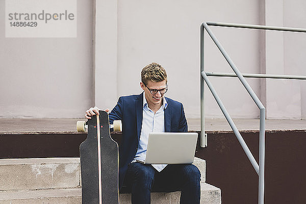 Lächelnder junger Geschäftsmann mit Skateboard sitzt draußen auf der Treppe und benutzt seinen Laptop